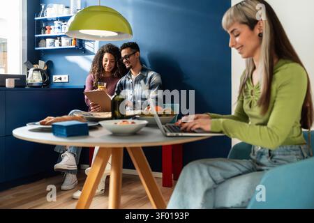 Multiethnic flatmates share a cozy kitchen at home, one working on a laptop while two friends read together and smile, showing modern roommate life an Stock Photo