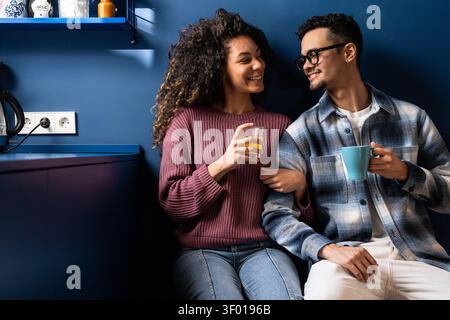 Happy latin couple sitting together in a modern blue kitchen, smiling and chatting while holding coffee and juice. Casual home lifestyle moment with w Stock Photo