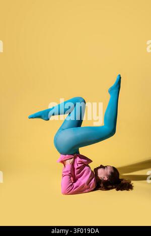 Young woman dancer performs a contemporary pose on the studio floor, wearing bright blue tights and a pink sweatshirt, set against a bold yellow backg Stock Photo
