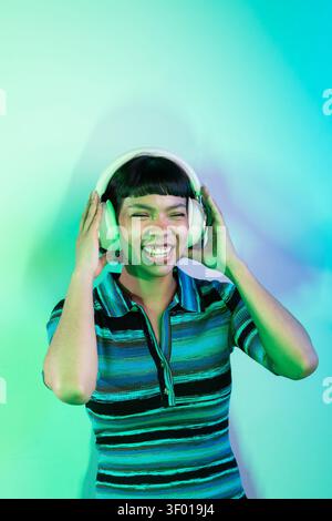 Smiling young latin woman wearing large headphones and a striped shirt, listening to music and laughing against a blue green neon gradient background Stock Photo