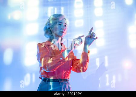 Futuristic senior Asian woman interacts with a holographic touchscreen interface, pointing at floating data in a neon-lit digital environment, showcas Stock Photo