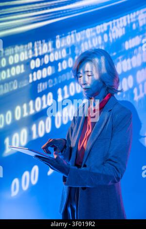Futuristic senior asian woman in a blazer uses a tablet under blue neon light, with binary code and digital data projected behind her, symbolizing AI, Stock Photo