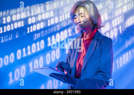 Futuristic senior Asian woman in a blazer uses a tablet under blue neon light, with binary code and digital data projection behind her, symbolizing AI Stock Photo