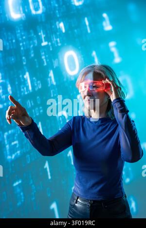 Futuristic senior Asian woman in blue top uses red AR glasses, pointing at glowing digital numbers and holographic interface, exploring AI, cybersecur Stock Photo