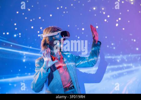 Futuristic senior Asian woman wearing a VR headset, reaching out with both hands in a glowing neon space, exploring immersive virtual reality and digi Stock Photo