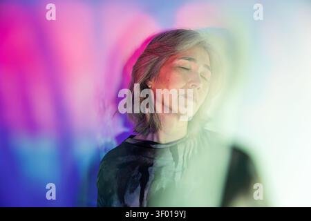Futuristic senior Asian woman portrait with closed eyes in neon pink and blue light, soft mist and bokeh background, serene mood, modern concept for t Stock Photo