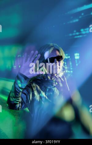 Futuristic senior Asian woman in reflective visor reaches toward a glowing holographic interface, surrounded by neon green and blue light, suggesting Stock Photo