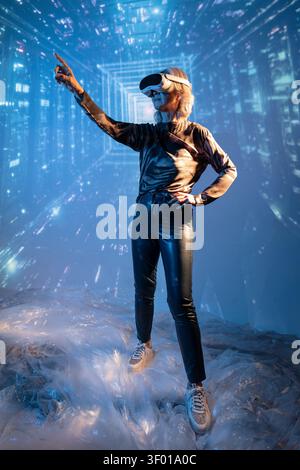 Futuristic senior Asian woman wearing a VR headset and black outfit, standing in a glowing digital tunnel and pointing forward, exploring immersive te Stock Photo