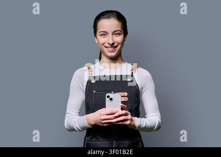 Confident young female in apron using smartphone posing on gray background Stock Photo