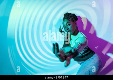Afro woman dancer striking a dynamic pose in neon blue and magenta studio lighting, with spiral projection background and strong shadow, modern urban Stock Photo