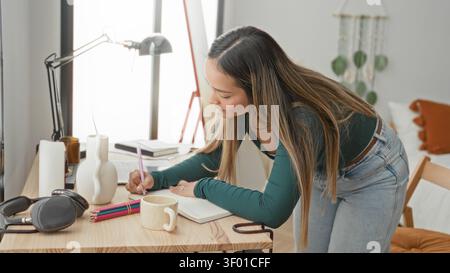 Woman leaning over desk writing in notebook with hand holding pen beside headphones and mug in a studio; concentration. Stock Photo