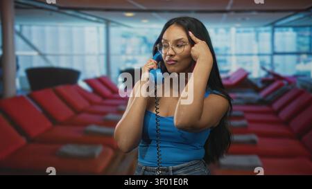Young hispanic woman holds vintage blue corded telephone handset to ear, finger to temple and hand on hip in an airport lounge with red seats; playful Stock Photo