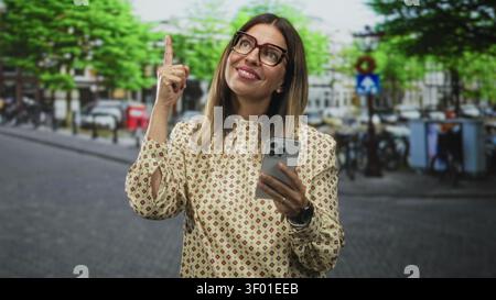 Woman holding smartphone points index finger up on a city street near bicycles and buildings; curiosity discovery inspiration. Stock Photo