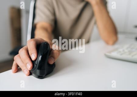 Close-up shot focuses on hand using black vertical ergonomic mouse. Person sits at white desk in modern office, preventing carpal tunnel syndrome Stock Photo
