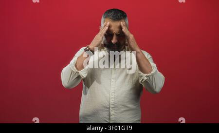Man pressing temples with hands, bearded middle age man wearing a white shirt in studio against a red wall; pain. Stock Photo