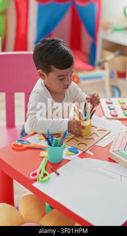 Boy with hand holding colored pencils and wooden shape puzzle on red table in building; curious concentration. Stock Photo