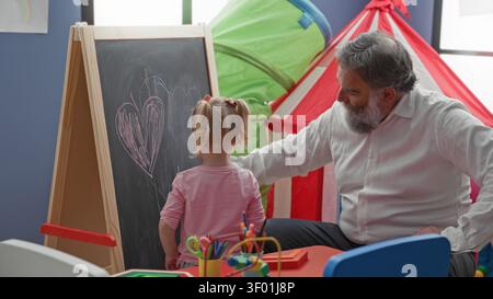 Man guiding girl's hand drawing heart on chalkboard in playroom building; nurturing learning moment. Stock Photo