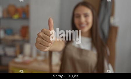 Young woman potter gives thumbs up showing hand and thumb in a pottery studio with shelves of clay pots and tools nearby; confidence creativity. Stock Photo