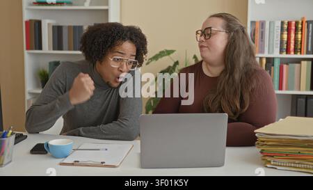 Man and woman leaning over laptop at desk with clipboard, coffee cup and stacked folders, man making a fist gesture while studying the screen in a bui Stock Photo