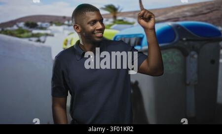 Young african man pointing finger upward beside blue recycling bins on street, smiling and gesturing; confidence optimism. Stock Photo
