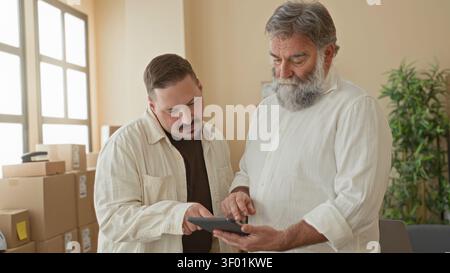 Two men point finger to tablet while checking stacked packages and discussing parcel labels in a building small business delivery office; collaboratio Stock Photo