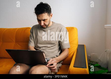 Concentrated young man in casual clothes working on laptop at home. Serious freelancer sitting on bright yellow sofa, focused on screen. Remote work Stock Photo