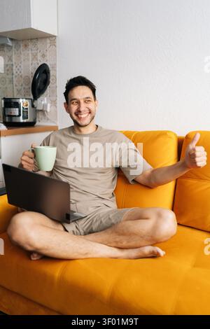 Happy young man works remotely on laptop from comfortable orange sofa. He smiles, drinks coffee, and shows thumbs up, expressing success and Stock Photo