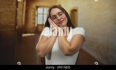 Woman in white tee wearing glasses, hands clasped near cheek with slight smile in old building interior by window; restful serenity. Stock Photo