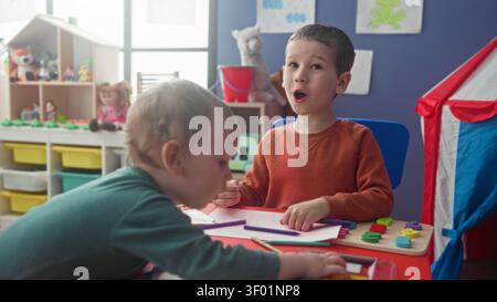 Boys drawing and reaching for colored pencils at a small table, toddler leaning over with hand on paper, play tent and toy shelves in building; playfu Stock Photo