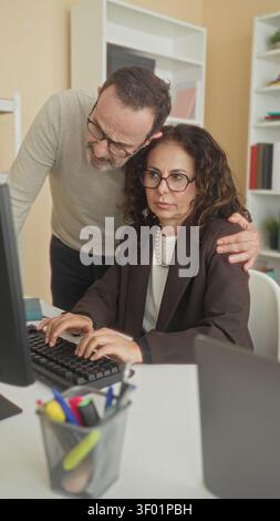 Woman typing on computer, middle aged man leaning over and placing hand on her shoulder while they review the screen in an office building; teamwork g Stock Photo