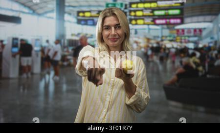 Young blonde woman holding lemon and pointing finger at camera in busy airport terminal near departure board and kiosks; playful invitation. Stock Photo