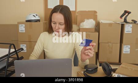 Woman holding creditcard and typing on laptop in packing warehouse among stacked parcels and barcode scanner; entrepreneurial focus. Stock Photo