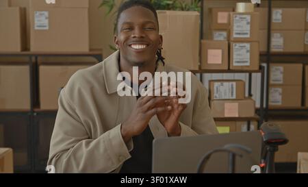 Man with packages clasping hands over laptop in a storage building with stacked boxes and scanner visible; small business pride calm. Stock Photo