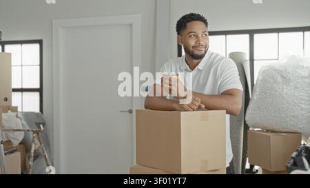 Man holding coffee cup and leaning on stacked cardboard boxes, relaxed smile while surrounded by bubble wrap and packing supplies in a building; new b Stock Photo