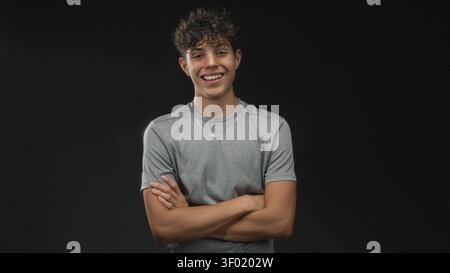 Boy wearing gray t shirt and silver necklace smiling with arms crossed in studio with black backdrop and soft lighting; youthful confidence. Stock Photo