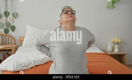 Woman middle age hoary short hair sitting on bed holds lower back, wincing and hunched forward on orange blanket with pillow and bedside table in a bu Stock Photo