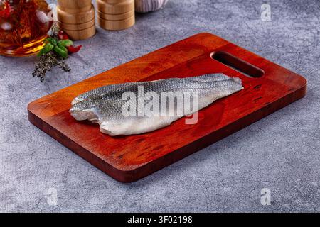 Fresh seabass fillet on rich red wood cutting board. Gray textured surface with soft lighting. Background includes garlic, herbs, oil, wooden grinders Stock Photo