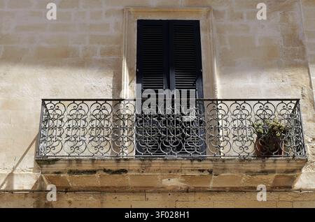 Detail of a vintage building facade featuring a decorative iron balcony and closed window shutters on a textured stone wall. Italy, lecce, 18 july 202 Stock Photo