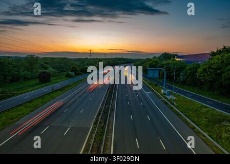 Long exposure of vehicle light trails on a multi-lane M4 motorway highway at sunset with an orange horizon sky and green trees, UK. Jct 13 Stock Photo