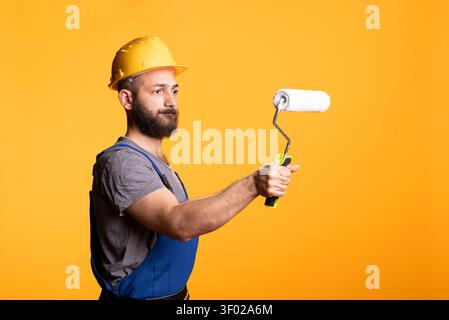 Painter in hardhat demonstrates roller for decorating or renovation jobs, presenting handyman service concept. Male construction worker stands in studio with painting tool against yellow backdrop Stock Photo