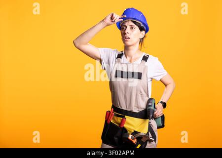 Female builder adjusts hardhat with puzzled expression, carrying power drill at waist. Renovation worker stands uncertainly in studio, dressed in overalls and tool belt against isolated background. Stock Photo