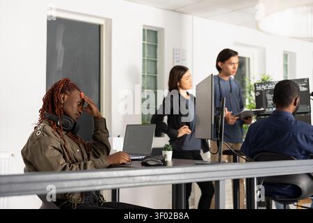 African American female coder concentrates at desk, contemplating software solutions with hand on forehead. Professional developer works on computer in collaborative startup office. Stock Photo