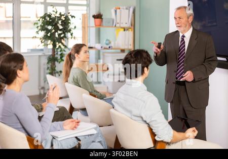 Senior teacher holding a remote control and speaking to student audience Professional senior teacher holding a remote control and speaking to student Stock Photo