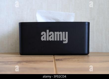 Front view of a minimalist black rectangular tissue box with a white paper napkin sticking out. Placed on a wooden table against a light neutral wall Stock Photo