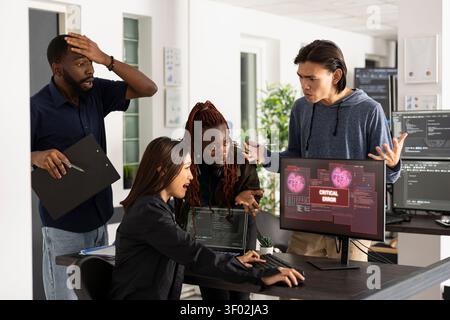 Professional IT team expresses shock and urgency while facing major cybersecurity attack. Frustrated developers looking at desktop monitor displaying red critical error warnings during data breach. Stock Photo