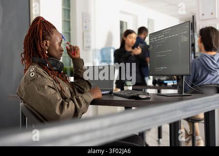 Black woman programmer sits at workstation, touching her forehead with pen in deep thought. Focused female developer analyzes data on screen as diverse colleagues collaborate in tech office. Stock Photo