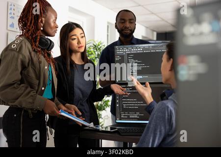 Manager addresses project delays as colleagues discuss coding progress and challenges. Team works together to solve issues, highlighting workplace communication and collaboration in web development. Stock Photo