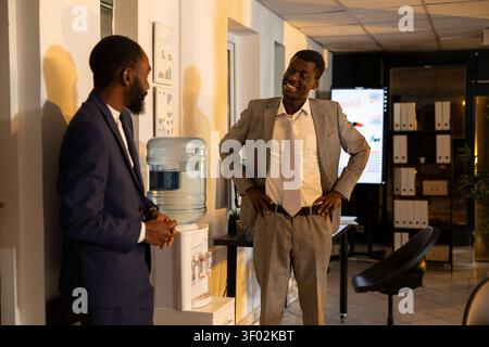 Black colleagues, chatting on break, bonding together, jokes at night, good friends sharing small talk and hanging out as a pastime after hours. Two office workers relaxing and socializing. Stock Photo