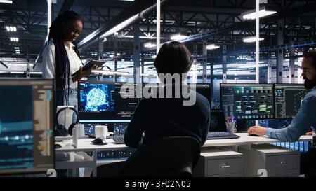 Technicians in high tech high tech server room cooperate on neural network software development, while IT staff members talking, implementing deep learning technology working with computer. Stock Photo