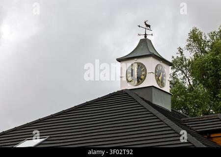 A white-painted clock tower with black and gold clock faces and a copper stag weathervane sits on the grey tiled roof of a village store building agai Stock Photo